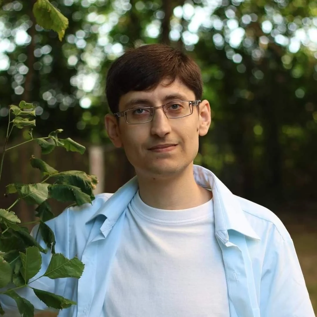 a person with glasses and a tshirt and long sleeve button shirt, with trees in foreground and background