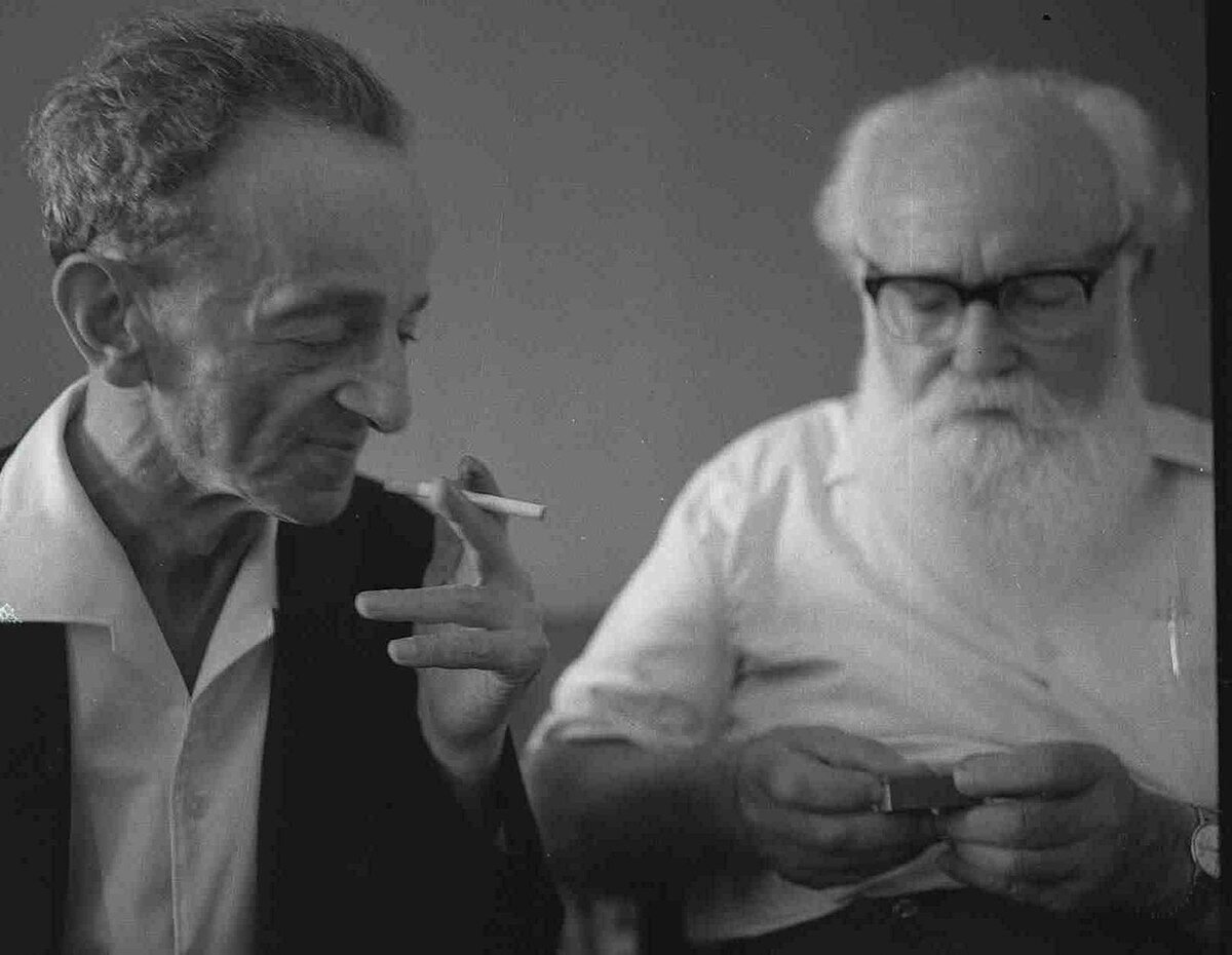 a black and white photo of Itzik Manger holding a cigarette and Yaakov Zerubavel is opening a box of matches to light it