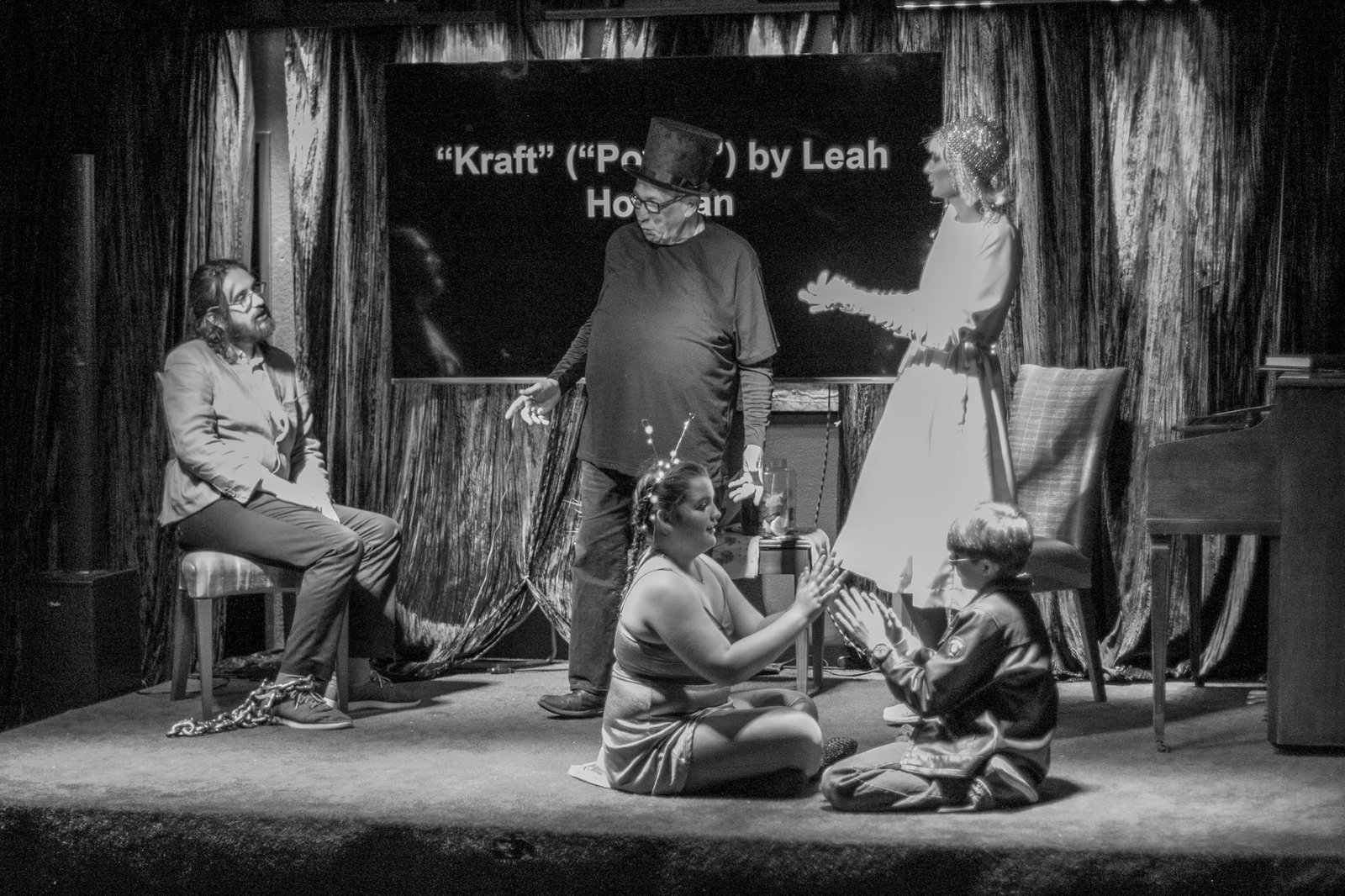 a black and white photo of 5 actors on stage in costume, dampf (steam) and elektri (electricity) are standing and speaking, shklaf (slave) is sitting on a chair and listening, and radio and airplane are sitting on the floor, playing pat-a-cake.