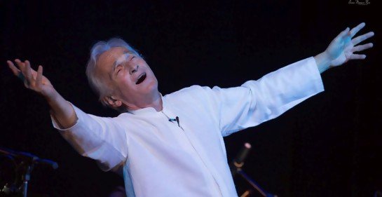 an actor with white hair and shirt spreading his arms wide, mouth open, head tilted back