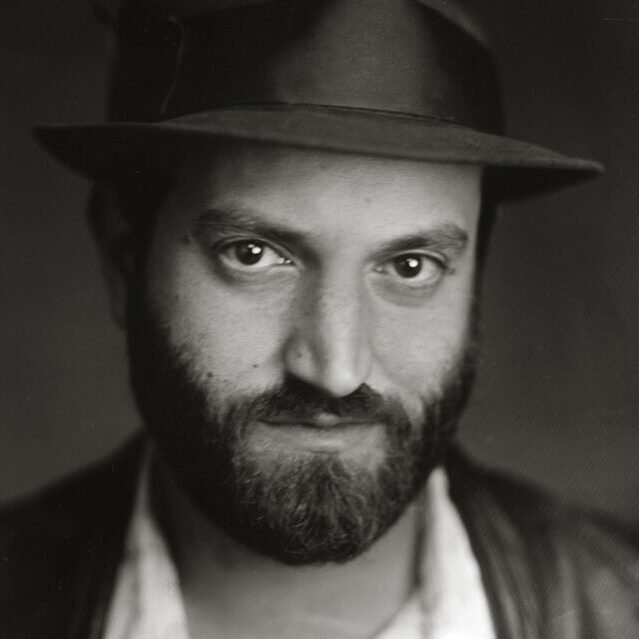 A black and white photograph of a man with a beard, hat and leather jacket.