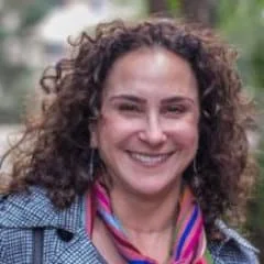 a photo of Elizabeth Scwartz, with a colorful scarf, and a jacket, smiling, natural background