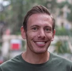 a person with short hair smiling, a street with trees in background