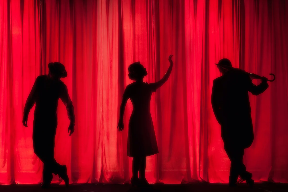 sillhouettes of three performers on stage, with a red curtain behind them